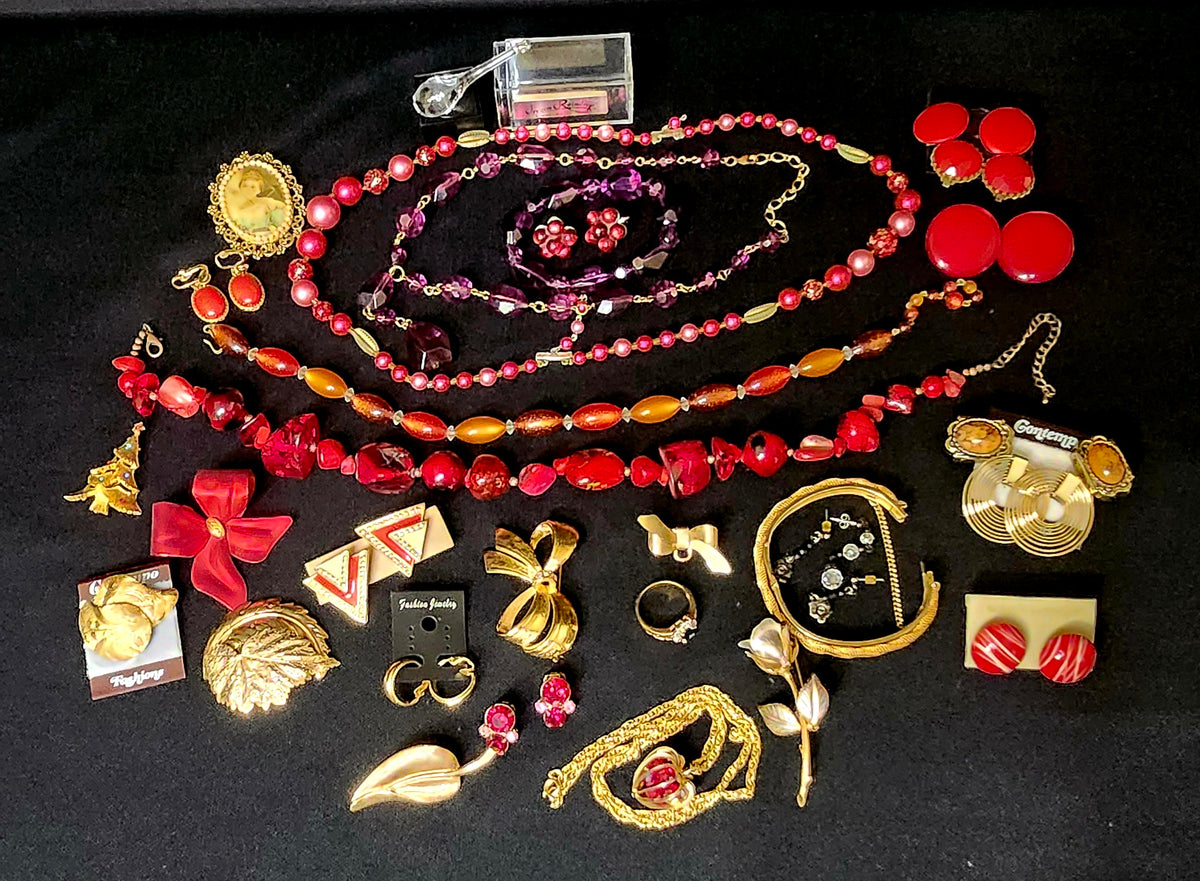 A collection of vintage jewelry and accessories arranged on a black background, including red and pink beaded necklaces, gold-tone earrings, bracelets, rings, and brooches, with some items still in their original retail packaging.