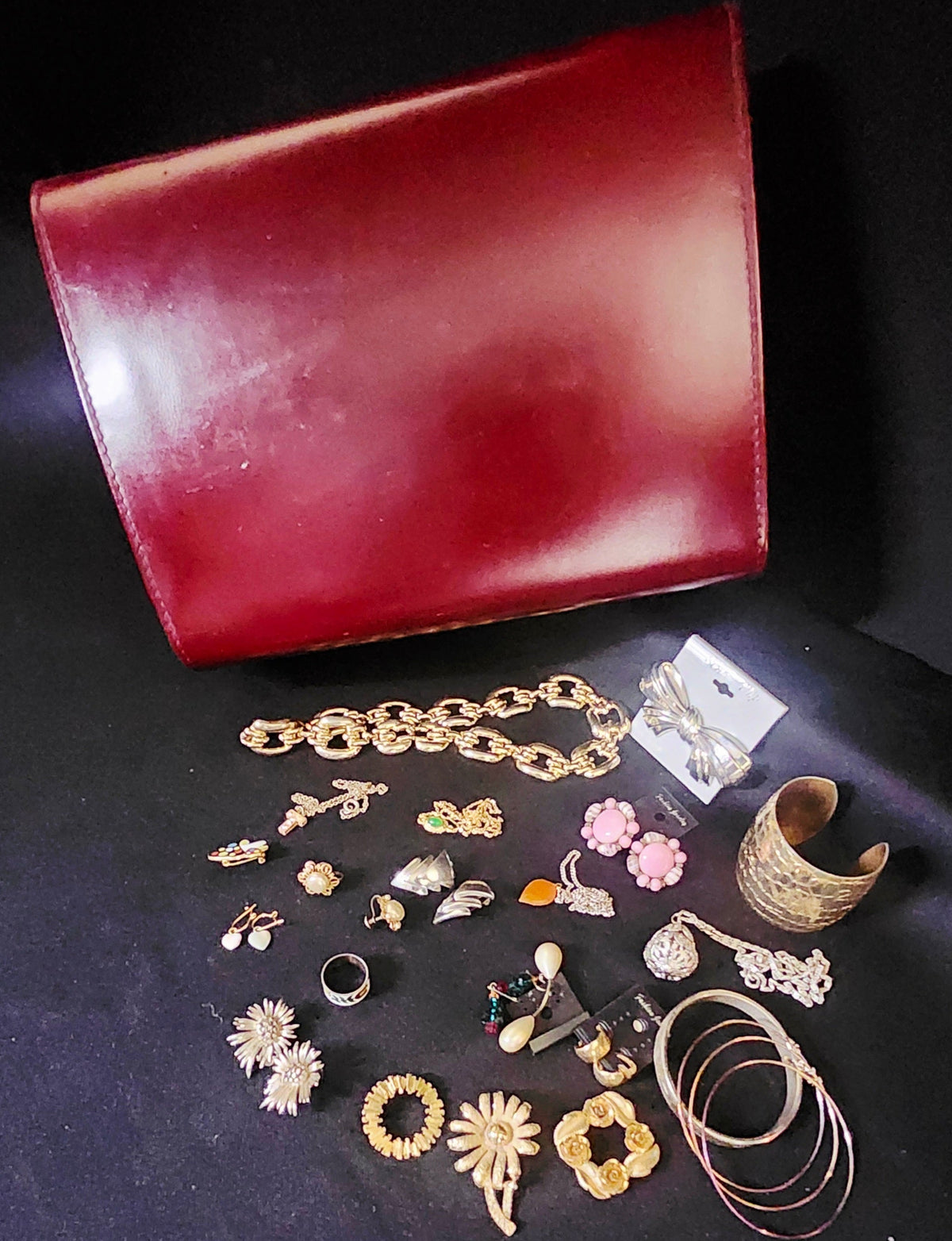 A collection of assorted vintage jewelry and accessories displayed on a black background, including a large red leather clutch, gold chain, earrings, rings, brooches, bracelets, and a gold cuff.