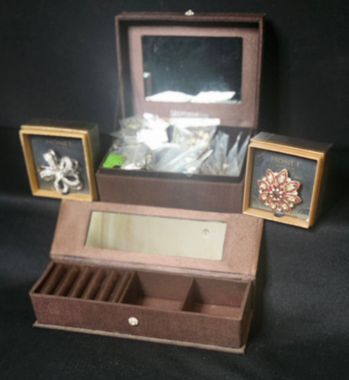 A brown velvet jewelry organizer box with an open top mirror, a lower compartment with dividers, and two small side frames holding brooches, photographed against a black background.