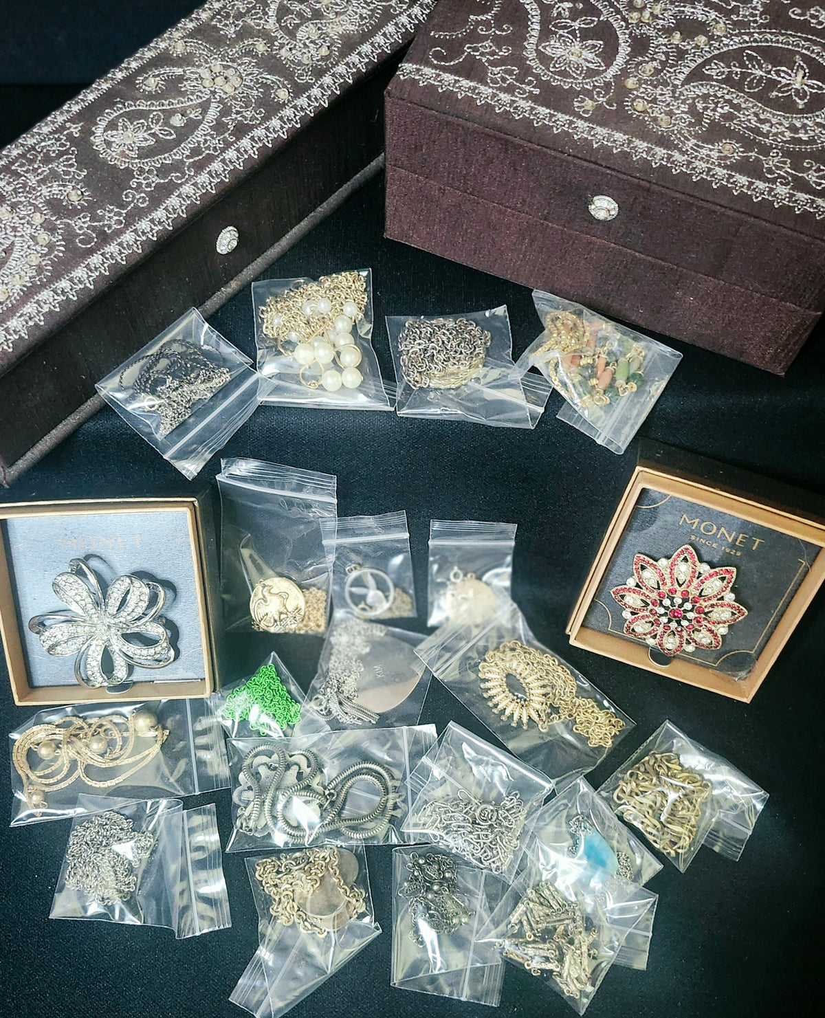 An assortment of jewelry pieces, including earrings and brooches, displayed in clear plastic bags and two decorative boxes on a black surface. The jewelry features gold, silver, and white metallic tones with some colorful accents.