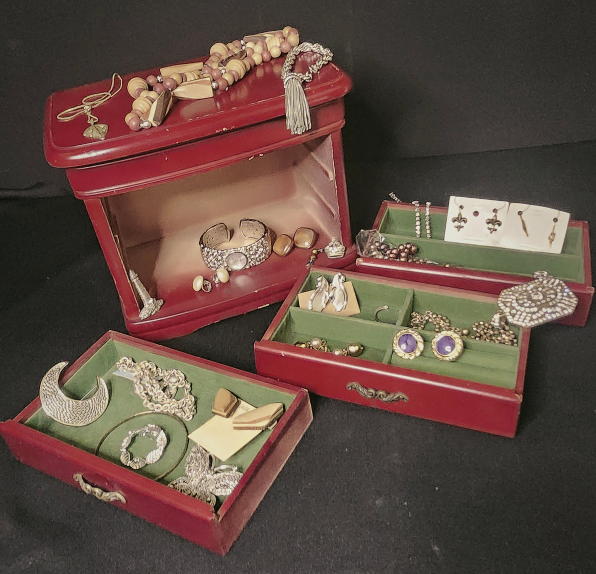 A set of vintage red wooden jewelry boxes with green velvet compartments, filled with assorted silver-toned jewelry including necklaces, watch, and earrings, displayed on a black background.