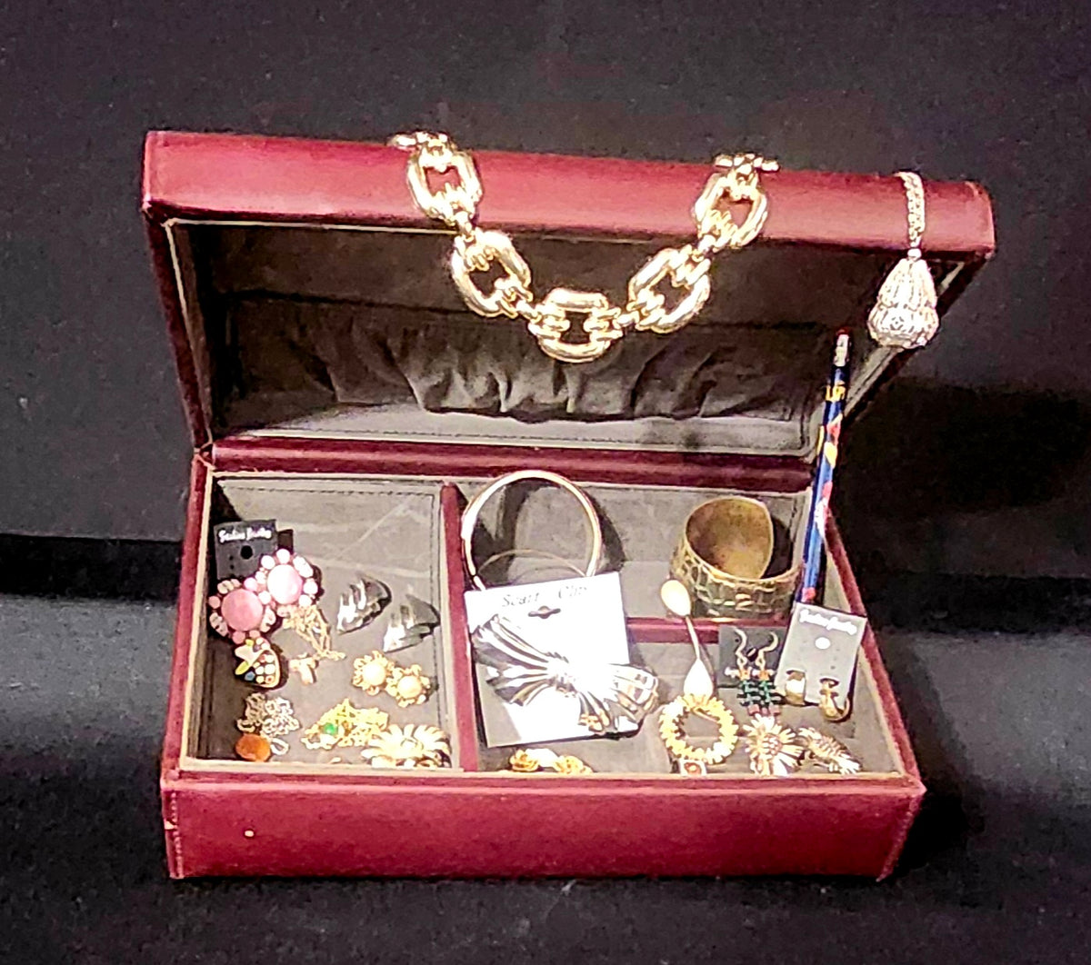 A vintage maroon leather jewelry box with its lid open, displaying a collection of assorted vintage jewelry including gold chain, silver rings, earrings, pendants, and a bracelet.