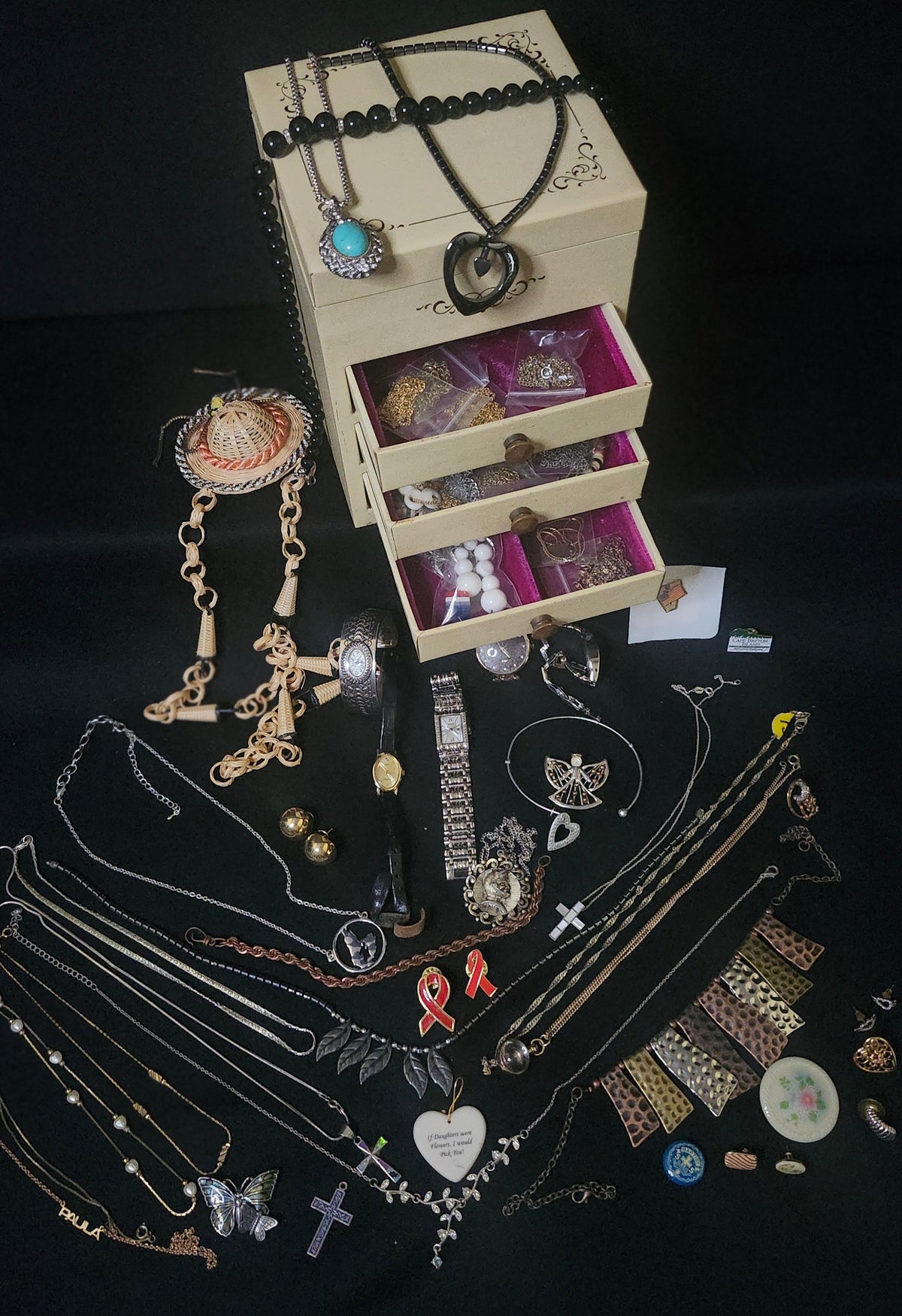 A collection of assorted jewelry pieces, primarily necklaces and bracelets, displayed on a black background alongside a cream-colored jewelry box with three open drawers.
