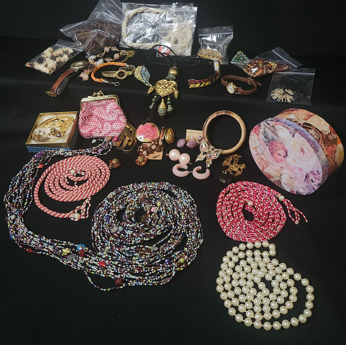 A collection of assorted vintage jewelry and accessories arranged on a black background, including multiple beaded necklaces, bracelets, earrings, a clutch purse, rings, and a floral-patterned box.