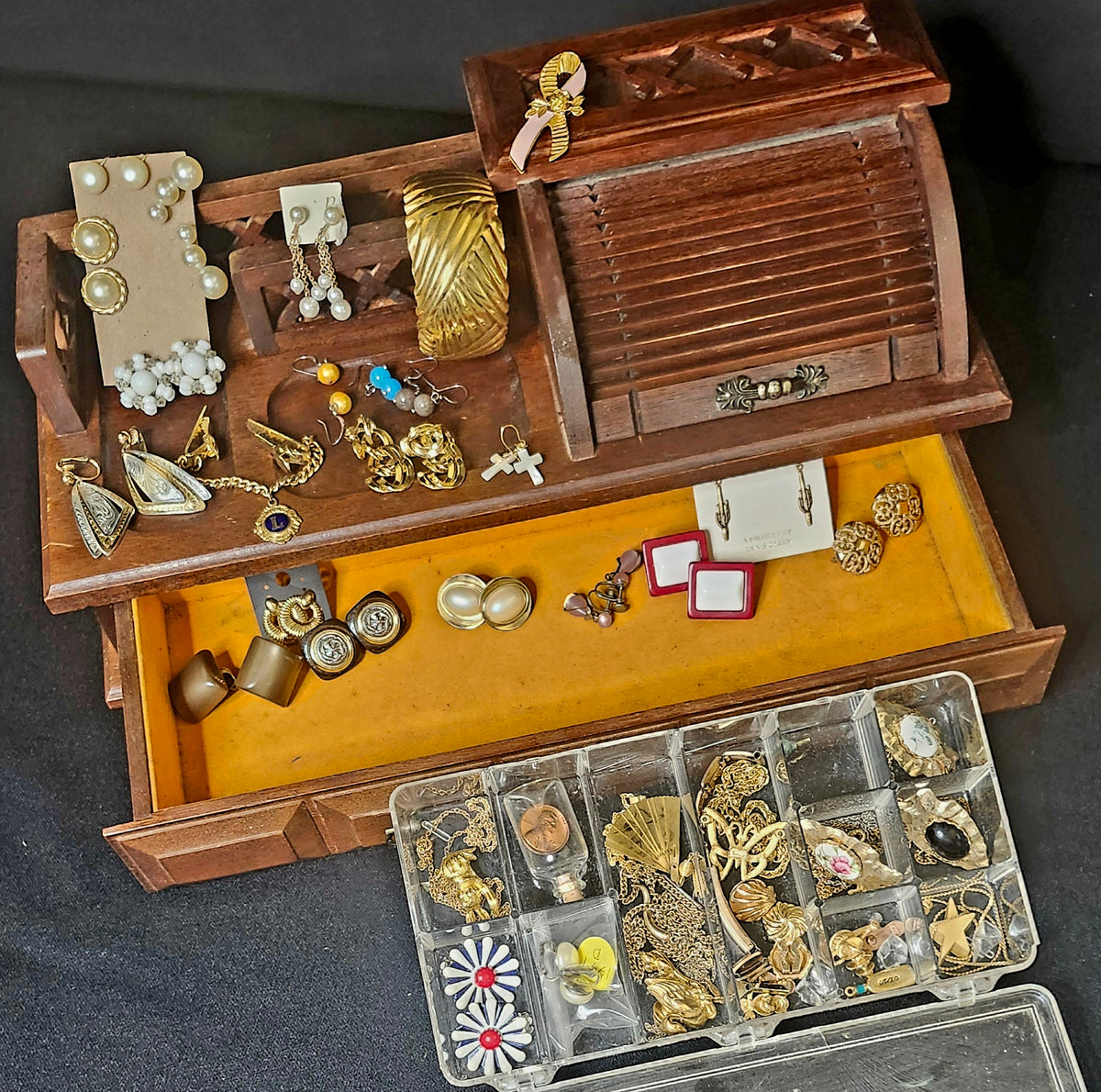 A wooden jewelry organizer with an open top compartment and a pulled-out drawer lined with yellow fabric, holding assorted vintage-style jewelry such as earrings, necklaces, rings, and brooches. A clear plastic compartment organizer sits in front on a dark fabric background.