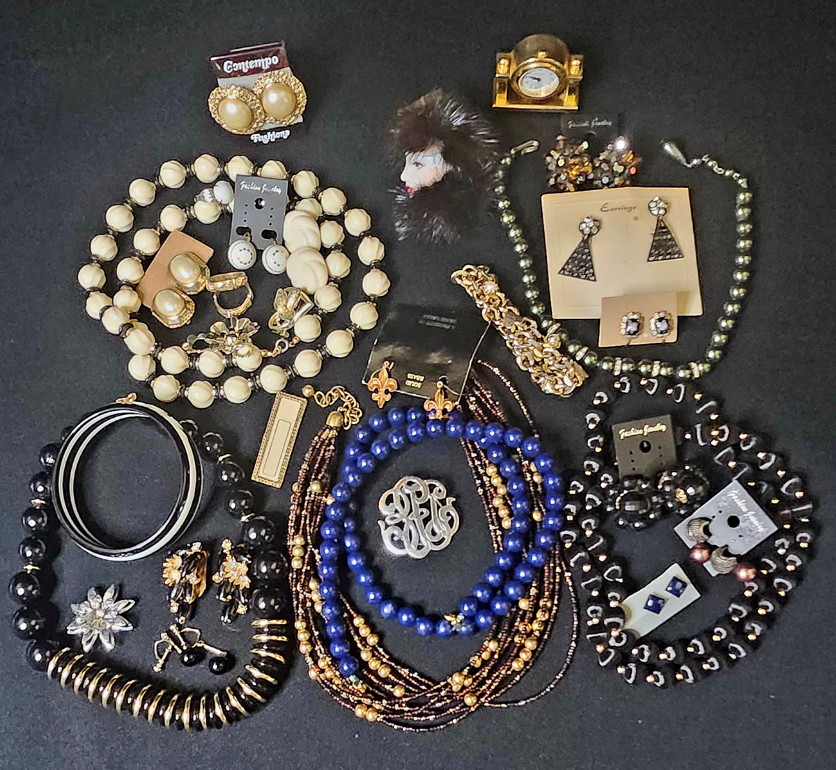 A flat lay of assorted jewelry and accessories arranged on a dark background, including multiple beaded necklaces, bracelets, earrings, rings, a watch and wrist bracelet.