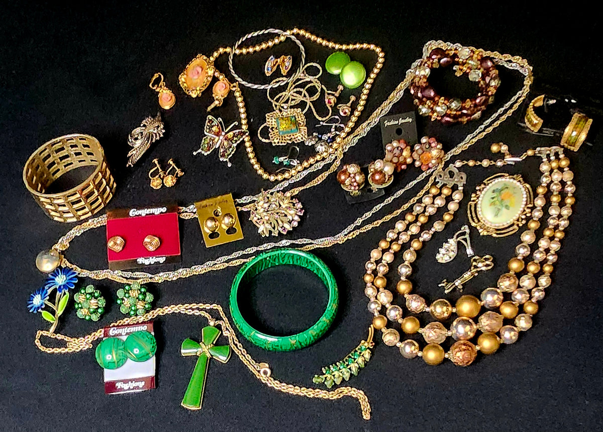 A collection of vintage costume jewelry laid out on a black background, including multiple gold-tone necklaces, beaded strands, a green bangle, earrings, a cross pendant.