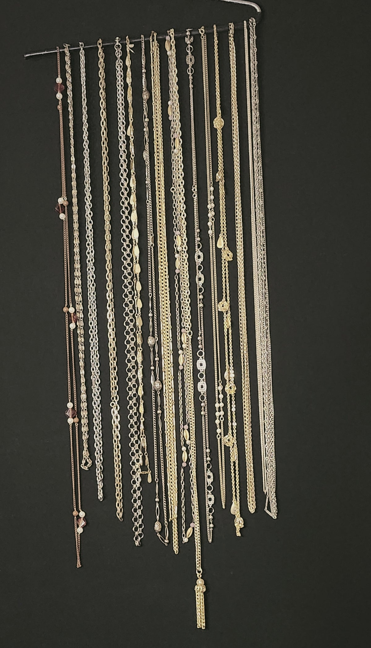 A collection of multiple long, delicate chains and necklaces in silver and gold tones hanging from a metal rod against a dark charcoal background.