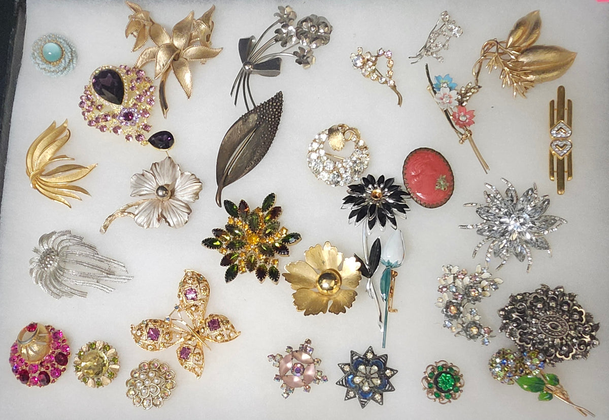 A collection of vintage floral and nature-themed brooches and pins arranged on a white background, featuring gold, silver, and multicolor designs with rhinestones and enamel.