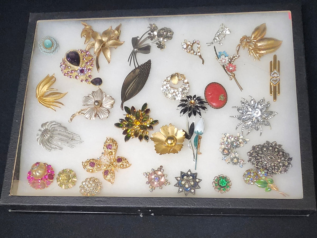 A collection of vintage-style brooches and pins arranged on a white display insert inside a black rectangular case, photographed from above on a dark surface.