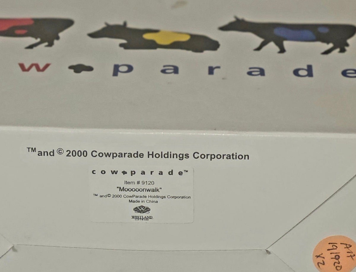 A close-up of a white product box for Cow Parade, showing printed branding, cow illustrations, a product label, and a barcode. A small orange photo sticker is affixed to the right side.
