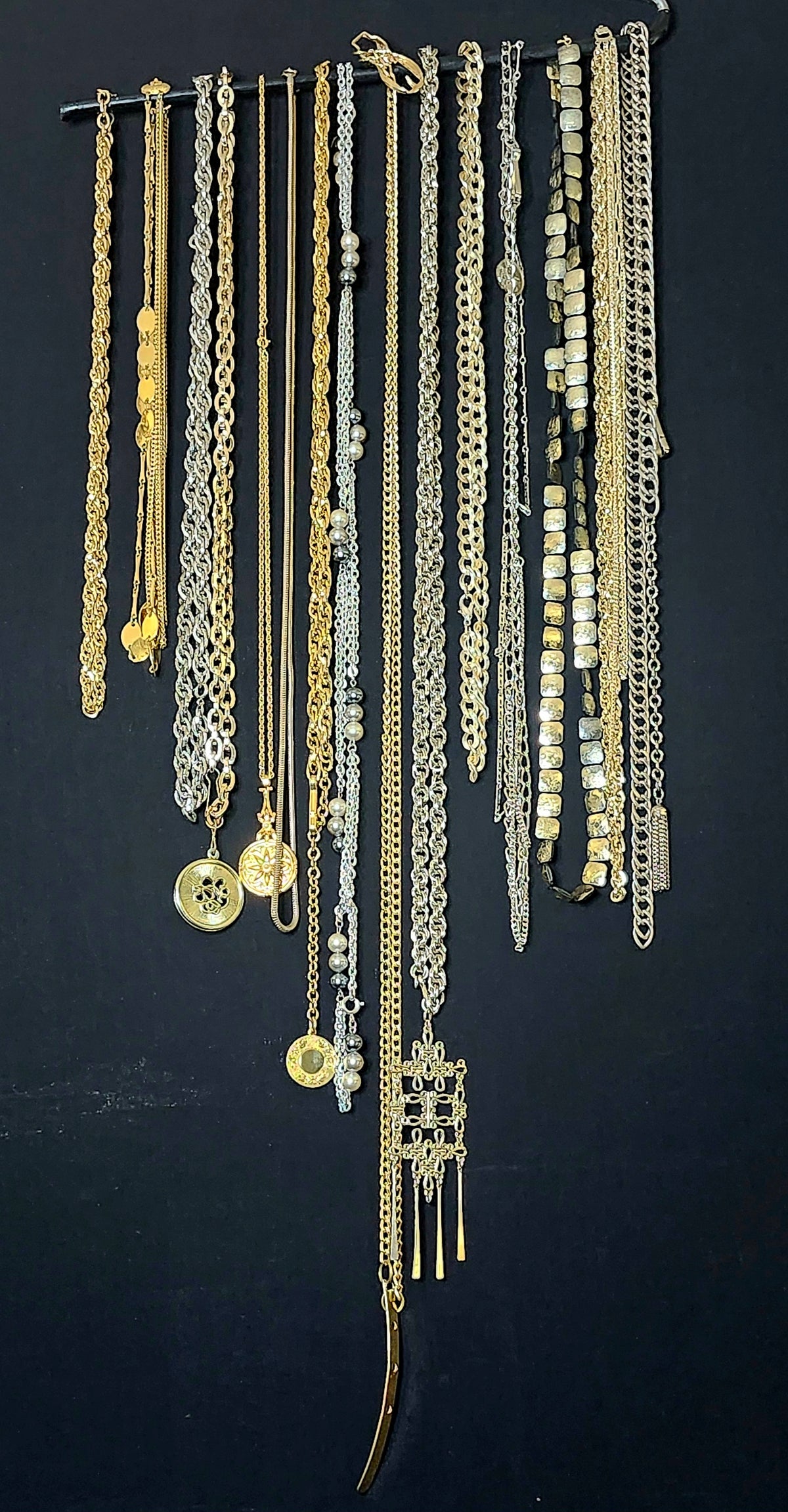 A collection of assorted gold and silver chain necklaces with various pendants, displayed hanging from a black rod against a dark background.