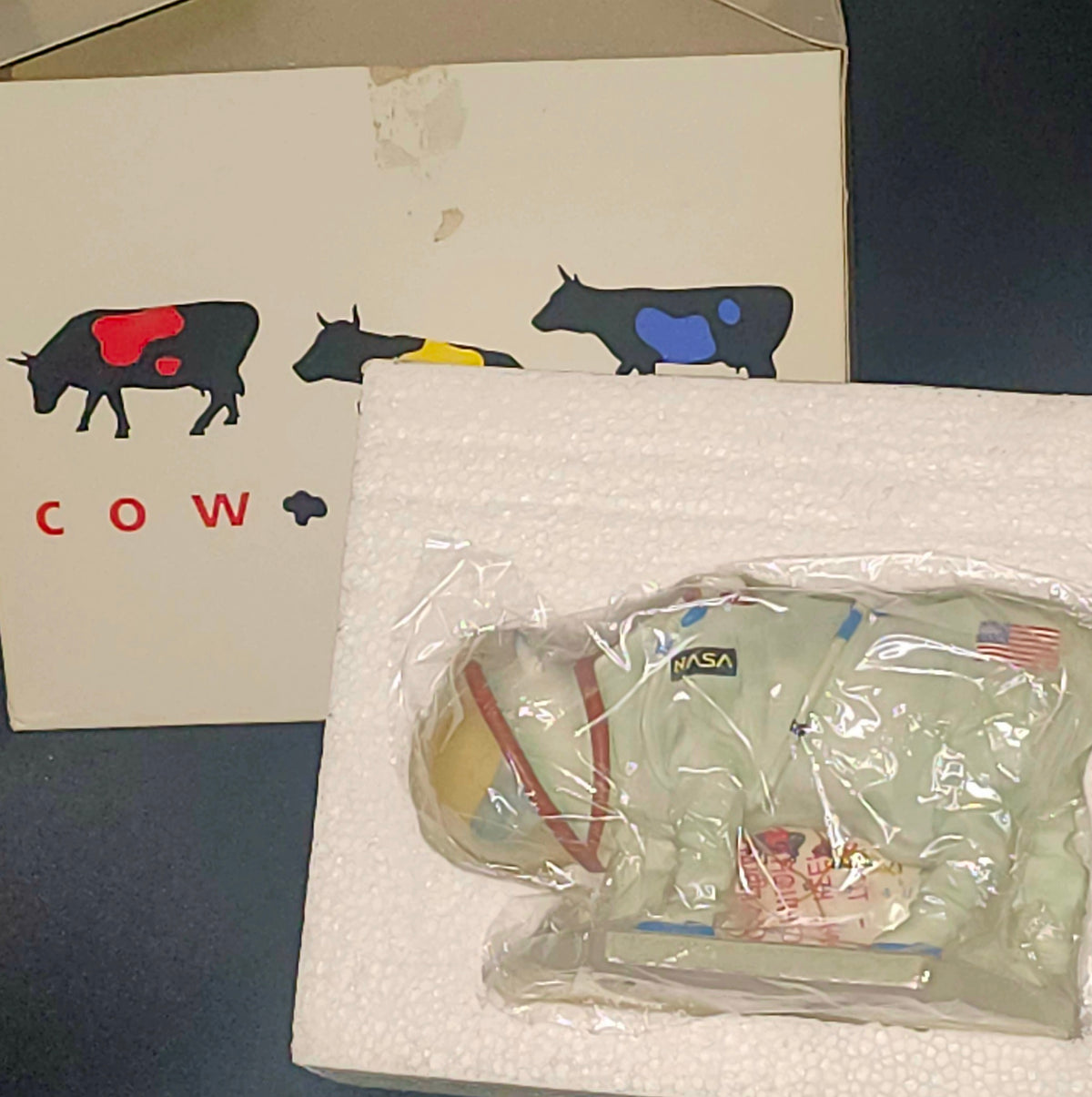 A cow-shaped astronaut figurine in a light green space suit, still sealed in clear plastic packaging and resting on white foam insert material, with its branded box visible in the background.